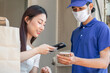 © KMPZZZ - Beautiful asian young woman, girl client use mobile phone scan accept delivery of baggage, cardboard box from delivery man due to lockdown, quarantine of covid-19 at entrance, door of her home, house.