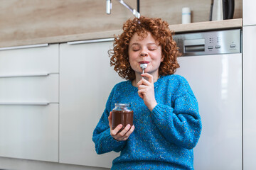 young woman eating chocolate from a jar while sitting on the wooden kitchen floor. Cute ginger girl indulging cheeky face eating chocolate spread from jar using spoon savoring every mouthful