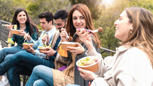 Gathering Of Young People Eating Take Away Organic Food In Plastic-free Bowls And Cutlery Together  In The Park. Focus On The Second From Right Smiling And Talking.