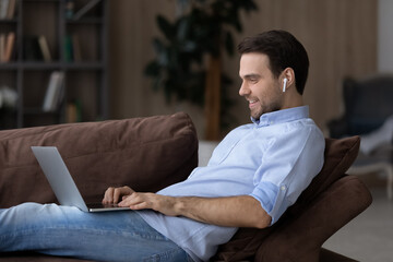 Wall Mural - smiling young caucasian man in wireless headphones sit on couch at home talk on video call on laptop