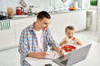 © insta_photos - Busy young parent father working or learning from home office using laptop computer sitting at kitchen table with cute kid child son eating toasts. Distance virtual work at homeoffice in family life.