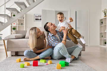 Happy young couple having fun with cute kid boy in modern living room. Parents playing with funny toddler child son, bonding together, enjoying love and happiness at home. Family mortgage concept.