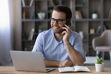 Canvas Print - Happy young Caucasian businessman sit at desk at home office work online on computer talk with client or customer on cellphone. Smiling man use laptop gadget at workplace have smartphone conversation.