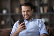 © fizkes - Smiling millennial Caucasian man look at cellphone screen speak talk on video call on gadget. Happy young male sit relax on the couch use smartphone texting or messaging online. Communication concept.