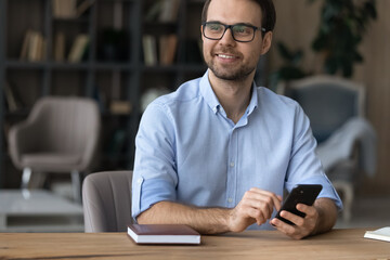 Canvas Print - Smiling young Caucasian man sit at desk at home use smartphone gadget look in window distance dreaming or planning. Happy male browse cellphone device think ponder of opportunities. Vision concept.