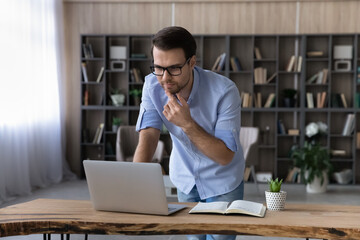 Poster - Pensive Caucasian man work online on laptop from home office make notes consult client or customer. Thoughtful young male look at computer screen make decision or plan, study distant on gadget.