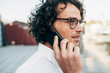 © iuricazac - Closeup side view of a businessman with curly hair talking with his friend on a mobile phone outdoor. Entrepreneur male wearing a white shirt and eyeglasses speaking on a cellphone.