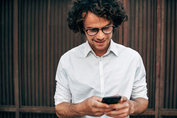 Wall Mural - Portrait of successful entrepreneur man in a white shirt with transparent eyeglasses messaging on a smartphone. The male has an online conference with colleagues to the lunch outside.