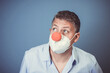 Middle aged man with nose mouth protection and red clown nose and blue shirt in front of blue background