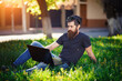 © endrews21 - Handsome bearded male software developer with a long mustache in a shirt and jeans sits with a laptop on the green grass in the park. Businessman holding an online conference in nature
