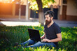 © endrews21 - An adult bearded male hacker with a long mustache in a shirt and jeans sits on green grass in the park and works on a laptop. Remote work via the Internet