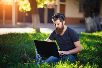 A bearded man with long dark hair with gray hair in a shirt and jeans sits on green grass in the park with a laptop. Internet surfing in free time