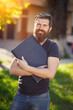 © endrews21 - Charismatic bearded male game developer with a long beard and dark gray hair in a shirt and jeans holds a laptop against the background of a green park in spring
