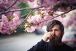 © endrews21 - A brutal bearded adult man with a long mustache and gray hair near in a dark sweater sniffs flowers near the cherry blossom trees in spring