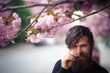 © endrews21 - A brutal bearded adult man with a long mustache and gray hair in a dark sweater sniffs flowers and laughs near cherry blossoms in spring