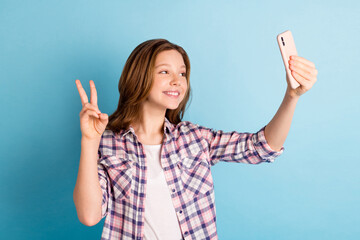 Poster - Photo of cute sweet school girl wear plaid shirt showing v-sign tacking selfie modern gadget isolated blue color background