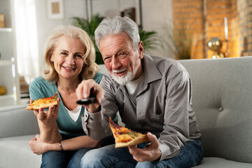 Wall Mural - Cheerful husband and wife sitting on sofa at home. Happy senior woman and man eating pizza while watching a tv