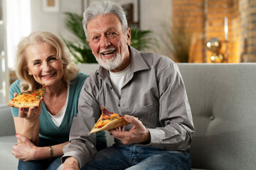 Wall Mural - Cheerful husband and wife sitting on sofa at home. Happy senior woman and man eating pizza while watching a tv