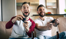 Excited Smiling Men Playing In Video Games On Tv At Home On The Couch. Friends With Joysticks Play Game With Happy Emotions On Faces