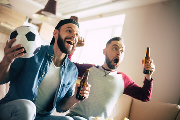 Funny excited hipster bearded friends with snacks and beers watching football on tv while sitting on the couch at home. Fans shouting due to winning of them team