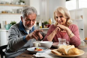 Wall Mural - Senior couple eating breakfast in the kitchen. Husband and wife talking and laughing while eating a sandwich..