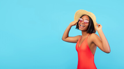 Happy black lady in bikini wearing sunglasses and straw hat, posing on blue background, banner design with free space