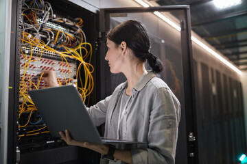Waist up of female network engineer connecting cables in server cabinet while working with supercomputer in data center, copy space