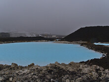 Pool With Blue Colored Thermal Water Between Lava Rocks With Steaming Svartsengi Power Station In Background Near Blue Lagoon, Grindavik, Reyjanes Peninsula, Iceland On Cloudy Day In Winter Season.