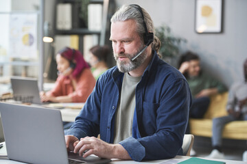 Wall Mural - Mature bearded man in headphones working on laptop at the table in call center