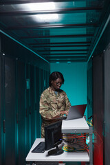 Vertical portrait of young African-American woman wearing military uniform using computer while setting up network in server room