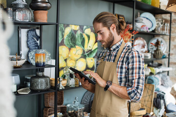 Wall Mural - Focused handsome bearded man using digital tablet for creating online catalogue of decor store. Positive seller in apron standing near shelves and looking at camera.