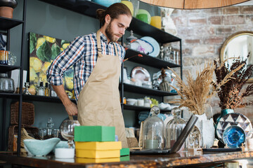 Wall Mural - Handsome caucasian man wearing beige apron working on modern laptop at decor store. Qualified seller creating online catalogue with available goods at shop.