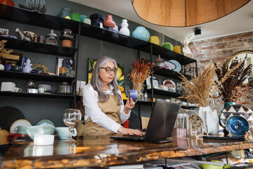 Wall Mural - Experienced saleswoman with grey hair doing inventory at decor store. Charming lady wering eyeglasses, white shirt and beige apron. Usage of modern laptop at work.