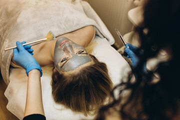 Professional beautician is applying healthy cream on female facial skin with concentration. Serene young woman is lying on table and enjoying treatment