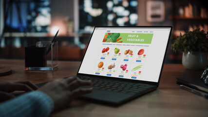 Wall Mural - Close Up on Hands of a Female Using Laptop Computer with Groceries Delivery Web Page to Order Fresh Vegetables for Dinner. Office Studio Apartment in the Evening with Big Windows.