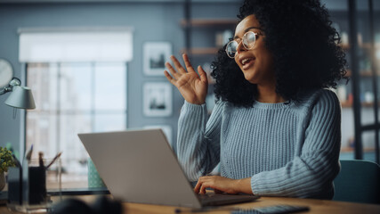 Beautiful Authentic Latina Female with Afro Hair Sitting at a Desk in a Cozy Living Room and Using Laptop Computer at Home. She's Browsing the Internet and Checking Videos on Social Networks.