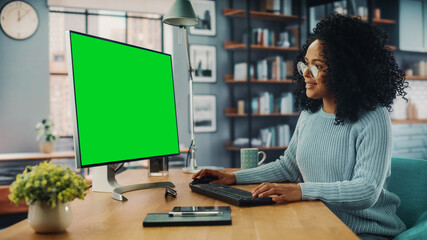 Latina Female Specialist Working on Desktop Computer with Green Screen Mock Up Display at Home Living Room while Sitting at a Table. Freelancer Female Chatting Over the Internet on Social Networks.
