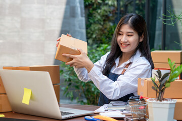 Young smiling beautiful owner asian woman freelance sme business online shopping working on laptop computer with parcel box on desk at home - SME business online and delivery concept