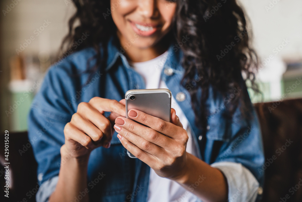 Cropped shot of an african-american young woman using smart phone at home. Smiling african american woman using smartphone at home, messaging or browsing social networks while relaxing on couch - obrazy, fototapety, plakaty 
