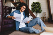 © InsideCreativeHouse - Positive young african-american woman reading documents and bills sitting on the floor at home. Accounting, taxes and finances concept - young african american woman with papers at home