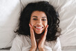 © InsideCreativeHouse - Close up portrait of a cheerful afro girl lying in her white bed in the morning. Top view of dark skinned woman , lies in bed with white bedclothes,touches both cheeks with hands. Afro lady in bedroom