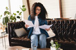 © InsideCreativeHouse - Clever 20s african woman sitting on the sofa and reading documents. Accounting, taxes and finances concept - young african american woman with papers at home