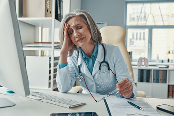 Tired mature female doctor in white lab coat keeping eyes closed while sitting in her office