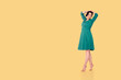 Woman stands on tiptoes holding her hands behind her head on pale orange-yellow background, dressed in dark cyan dress.Free space for your text.