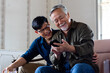 © Nutnutchar - Son cuddle dad while using mobile phone, smiling and laughing together. Old Asian man learning to use smartphone by young Asian man suggest him. Father's day and happy family concept.