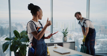 Cleaning services team. Young man using vacuum cleaner tidying office workspace. Female african american cleaning employee using tablet computer.