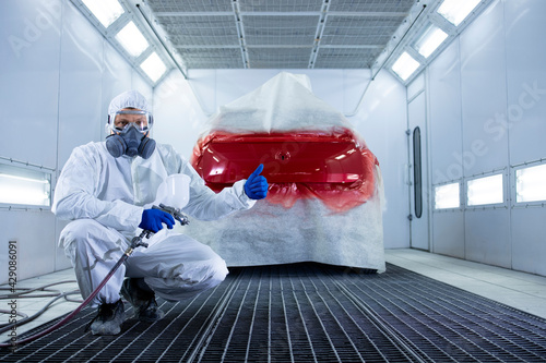 Portrait of professional car painter with protective clothing and mask holding thumbs up and standing by automobile in the painting chamber.