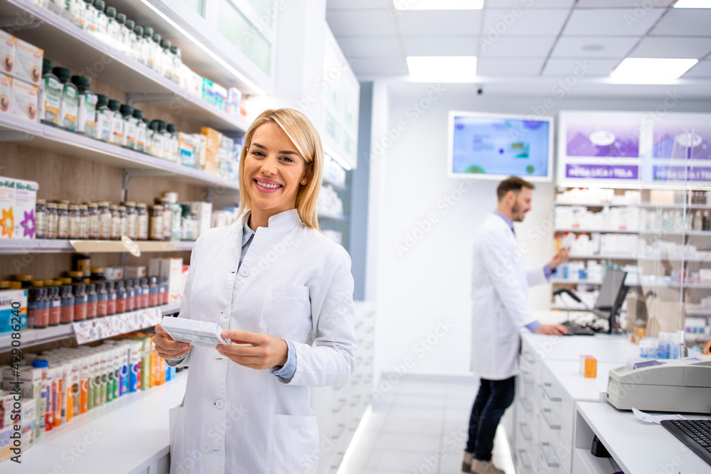 Portrait of beautiful female pharmacist standing in pharmacy store by the shelf with medicines. - obrazy, fototapety, plakaty 