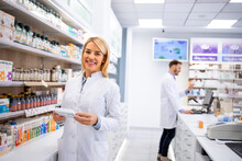 Portrait Of Beautiful Female Pharmacist Standing In Pharmacy Store By The Shelf With Medicines.