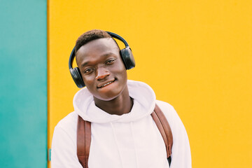 Wall Mural - Young afro American black man listening music with wireless headphones while wearing a white sweatshirt and a backpack looking to camera on a green and yellow wall background